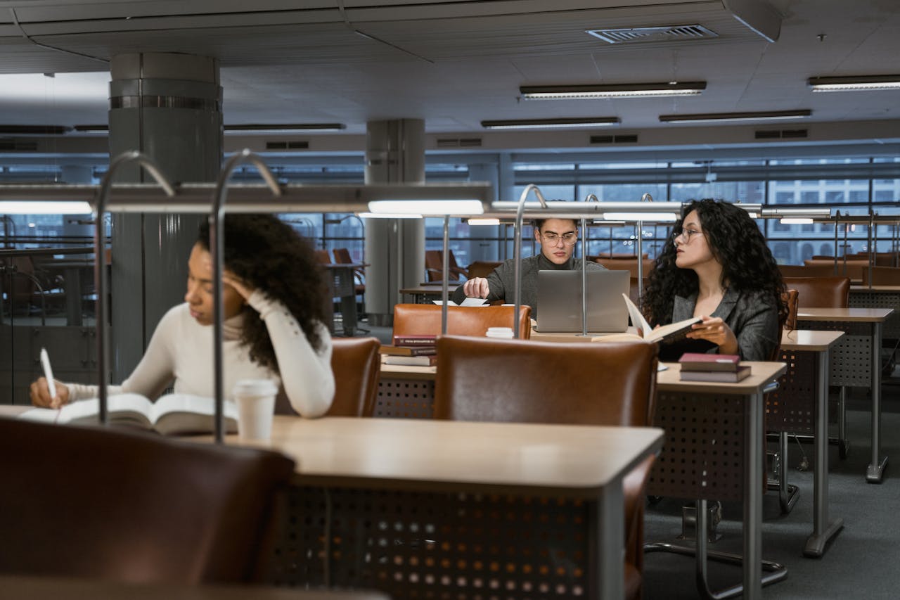 College students studying in a modern library setting, concentrating on books and laptops.