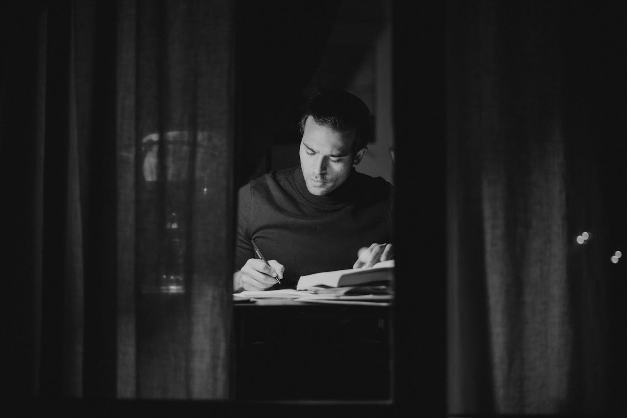 gallery-1 A man deeply focused on studying at night, captured in black and white from an indoor perspective.